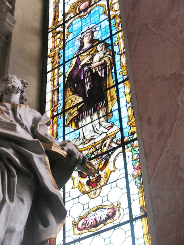 Saint Agnes stained-glass window in the Ursuline Church, Sibiu, with a marble saint’s statue in the foreground.