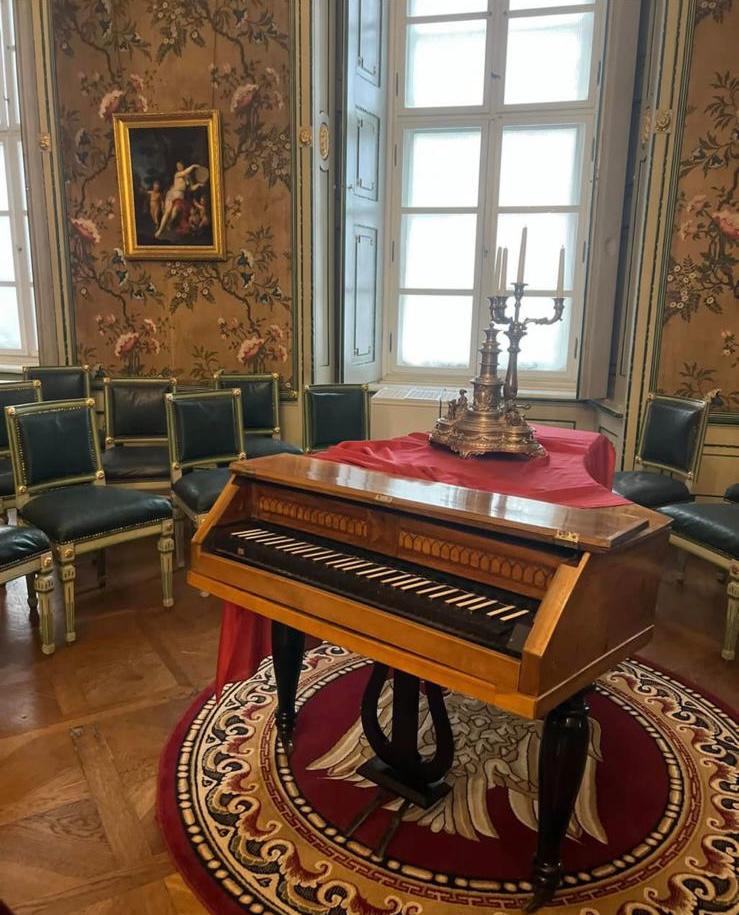 Brukenthal Palace salon—historic keyboard instrument, silver candelabrum on red drape, green neoclassical chairs, floral wall covering.