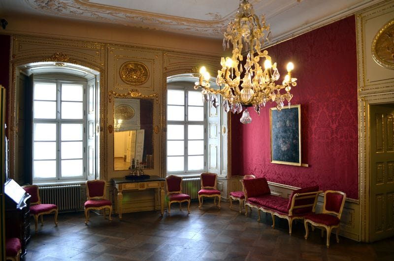 Brukenthal Palace salon—red damask walls, gilded panels and seating, crystal chandelier, tall windows with shutters.